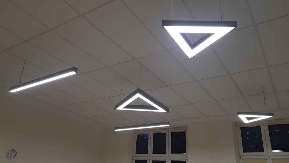 Nowoczesne oświetlenie sufitowe z lamp LED w kształcie trójkątów i prostokątów, zawieszone na cienkich linkach w pomieszczeniu z kasetonowym sufitem i oknami.