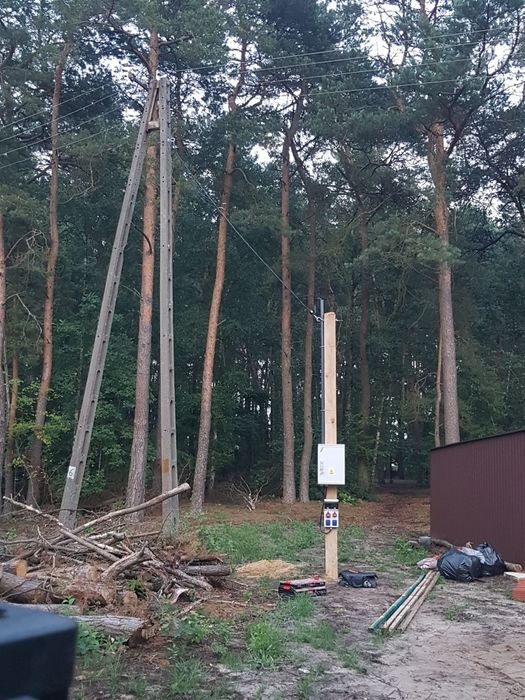 Nowy słup energetyczny z zamontowaną skrzynką rozdzielczą, widoczne narzędzia pracy i stary słup w tle lasu.