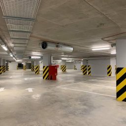 Palmil Sp. z o.o. - Wnętrze parkingu podziemnego z widoczną instalacją wentylacyjną pod sufitem, słupy pomalowane w czarno-żółte pasy.