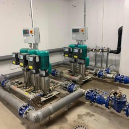 Palmil Sp. z o.o. - Zestaw czterech pomp hydroforowych z zielonymi silnikami, połączonych rurociągami z niebieskimi zaworami, zainstalowany w sterylnym pomieszczeniu technicznym.