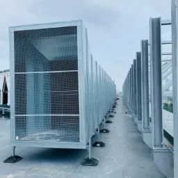 Szereg srebrnych, metalowych kanałów wentylacyjnych z siatką ochronną, ustawionych na dachu budynku, z widocznymi wspornikami i elementami mocującymi.