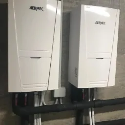 Dwa białe urządzenia Aermec zamontowane na szarej, betonowej ścianie, z widocznymi czarnymi rurami izolowanymi i białymi przewodami.