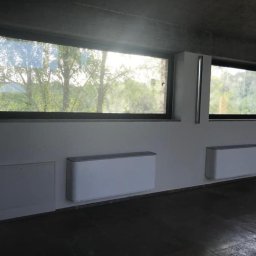 Palmil Sp. z o.o. - Dwa białe, poziome grzejniki ścienne zamontowane pod oknami z widokiem na zieleń, w pomieszczeniu z surowym betonowym sufitem i ciemną podłogą. Po lewej stronie widoczna skrzynka rewizyjna...