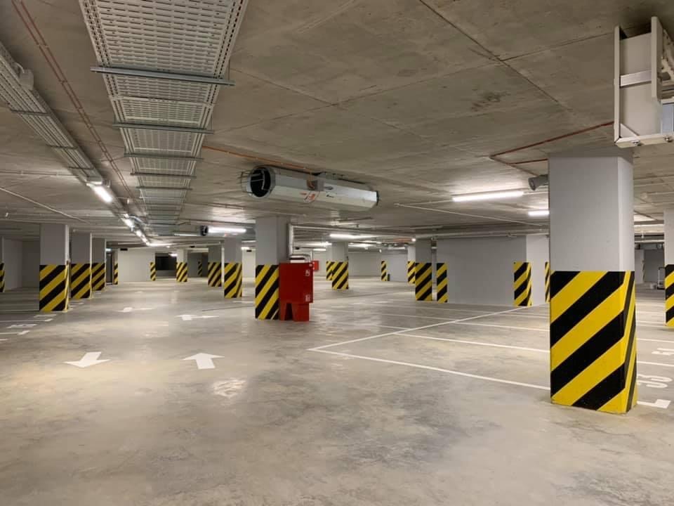 Wnętrze parkingu podziemnego z widoczną instalacją wentylacyjną pod sufitem, słupy pomalowane w czarno-żółte pasy.