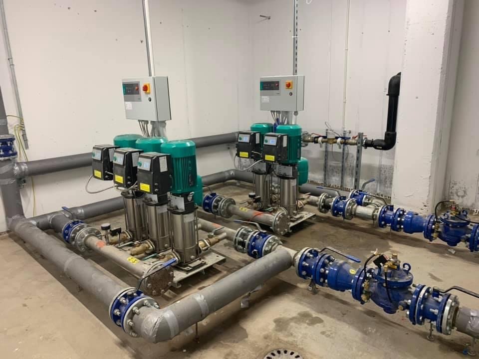 Zestaw czterech pomp hydroforowych z zielonymi silnikami, połączonych rurociągami z niebieskimi zaworami, zainstalowany w sterylnym pomieszczeniu technicznym.