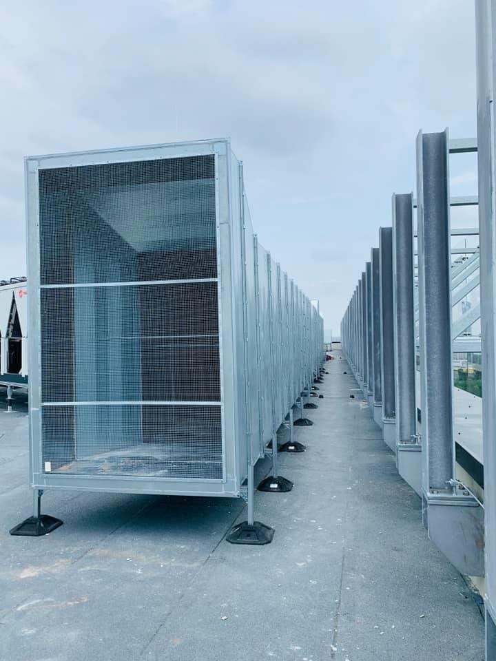 Szereg srebrnych, metalowych kanałów wentylacyjnych z siatką ochronną, ustawionych na dachu budynku, z widocznymi wspornikami i elementami mocującymi.