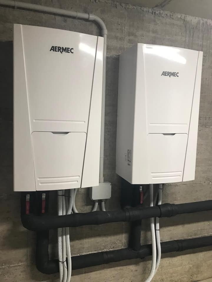 Dwa białe urządzenia Aermec zamontowane na szarej, betonowej ścianie, z widocznymi czarnymi rurami izolowanymi i białymi przewodami.