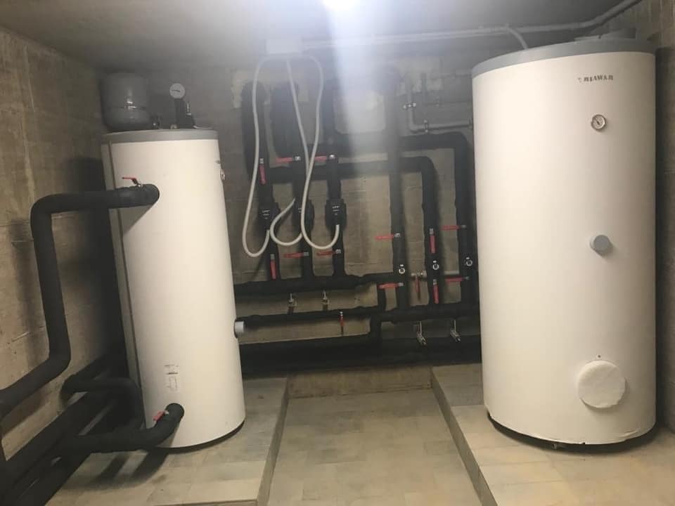 Dwa białe zbiorniki, prawdopodobnie na wodę, z czarnymi rurami i zaworami w piwnicy z betonowymi ścianami i podłogą wyłożoną płytkami. Jeden zbiornik ma manometr na górze i napis 'MEWAR' w prawym...