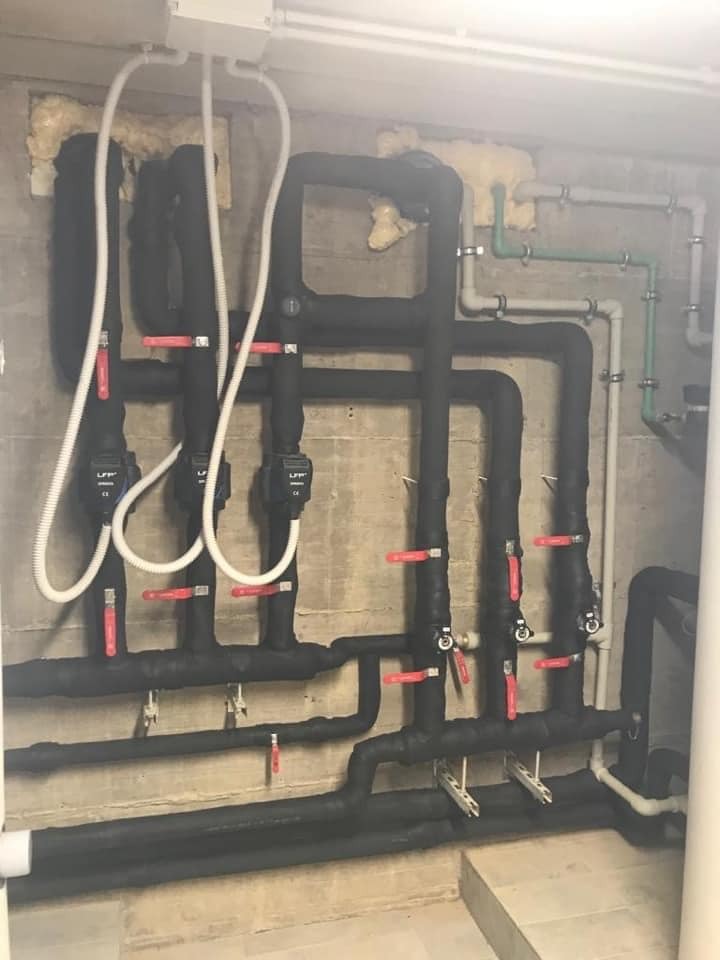 Złożony system rur grzewczych z czarną izolacją i czerwonymi zaworami, zamontowany na surowej betonowej ścianie w piwnicy, widoczne białe rury i elementy instalacji elektrycznej pod sufitem.