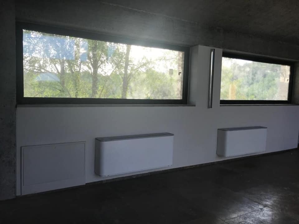 Dwa białe, poziome grzejniki ścienne zamontowane pod oknami z widokiem na zieleń, w pomieszczeniu z surowym betonowym sufitem i ciemną podłogą. Po lewej stronie widoczna skrzynka rewizyjna...