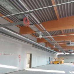 Wnętrze hali w Berlinie w trakcie prac instalacyjnych: widoczne kable, rury wentylacyjne pod sufitem z drewnianą konstrukcją, podnośniki koszowe i pracownik w oddali.