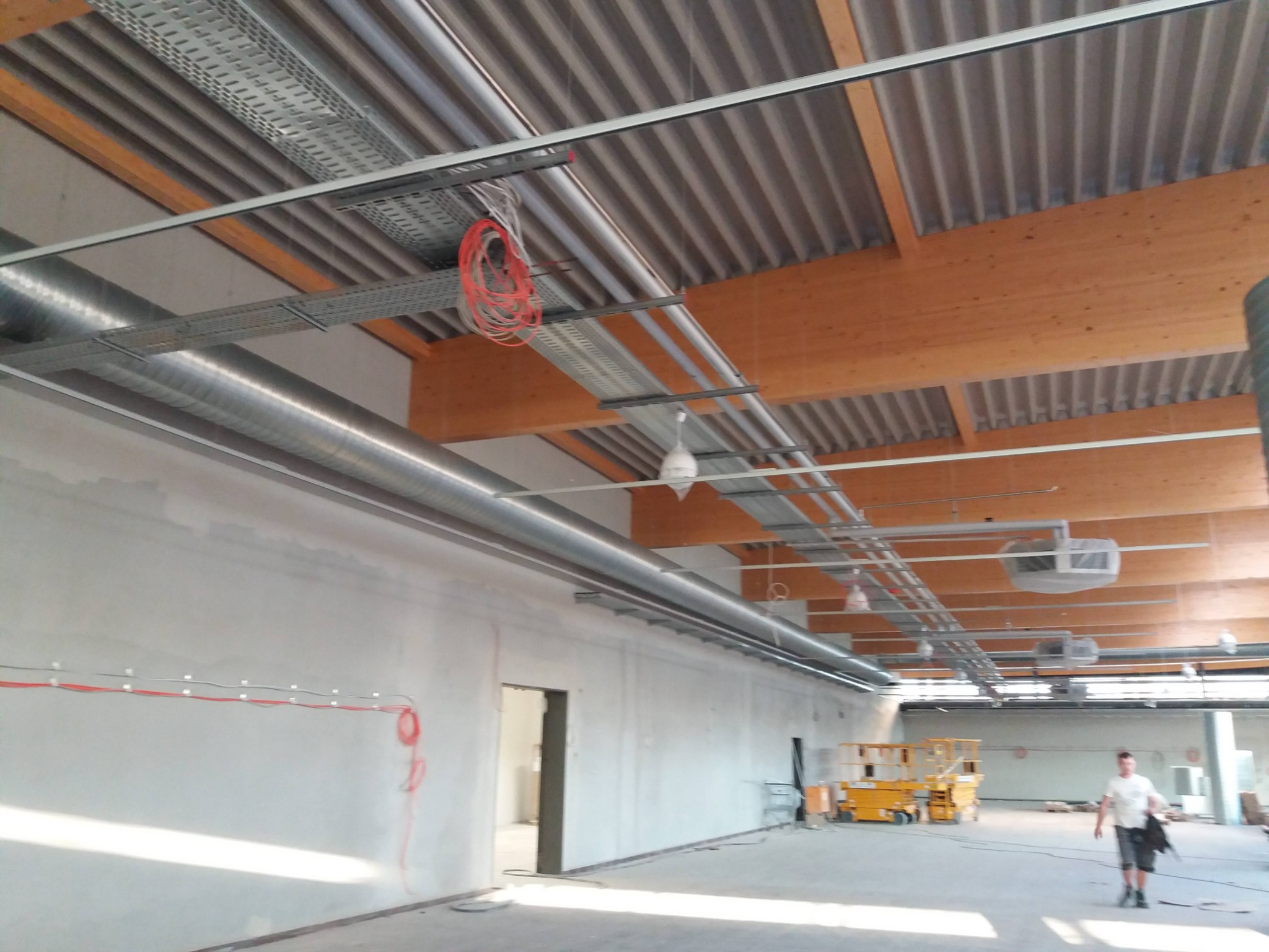 Wnętrze hali w Berlinie w trakcie prac instalacyjnych: widoczne kable, rury wentylacyjne pod sufitem z drewnianą konstrukcją, podnośniki koszowe i pracownik w oddali.