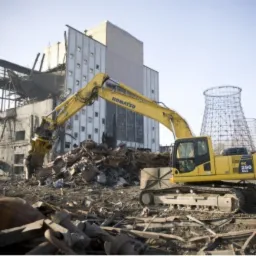 Żółta koparka Komatsu PC 290 NLC na tle gruzów i częściowo zburzonego budynku przemysłowego z widocznymi chłodniami kominowymi w tle.