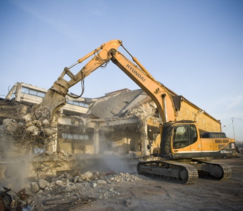 Żółta koparka Hyundai 320LC-9 z hydraulicznym młotem kruszy pozostałości budynku podczas rozbiórki w słoneczny dzień.