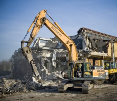 Żółta koparka Hyundai z hydraulicznym młotem kruszy betonową konstrukcję budynku podczas rozbiórki, widoczny operator w kabinie i druga maszyna w tle.
