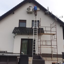 KLIMSPEC Przemysław Stróżewski - Instalator w niebieskiej kurtce i czerwonej czapce stoi na drabinie przy ścianie domu, montując jednostkę zewnętrzną klimatyzacji. Obok drabiny stoi rusztowanie. Na pierwszym planie widoczne meble...