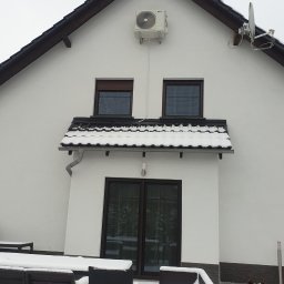 KLIMSPEC Przemysław Stróżewski - Biały dom jednorodzinny z zamontowaną klimatyzacją na ścianie szczytowej, antena satelitarna obok, widoczne meble ogrodowe pokryte śniegiem oraz owczarek niemiecki.