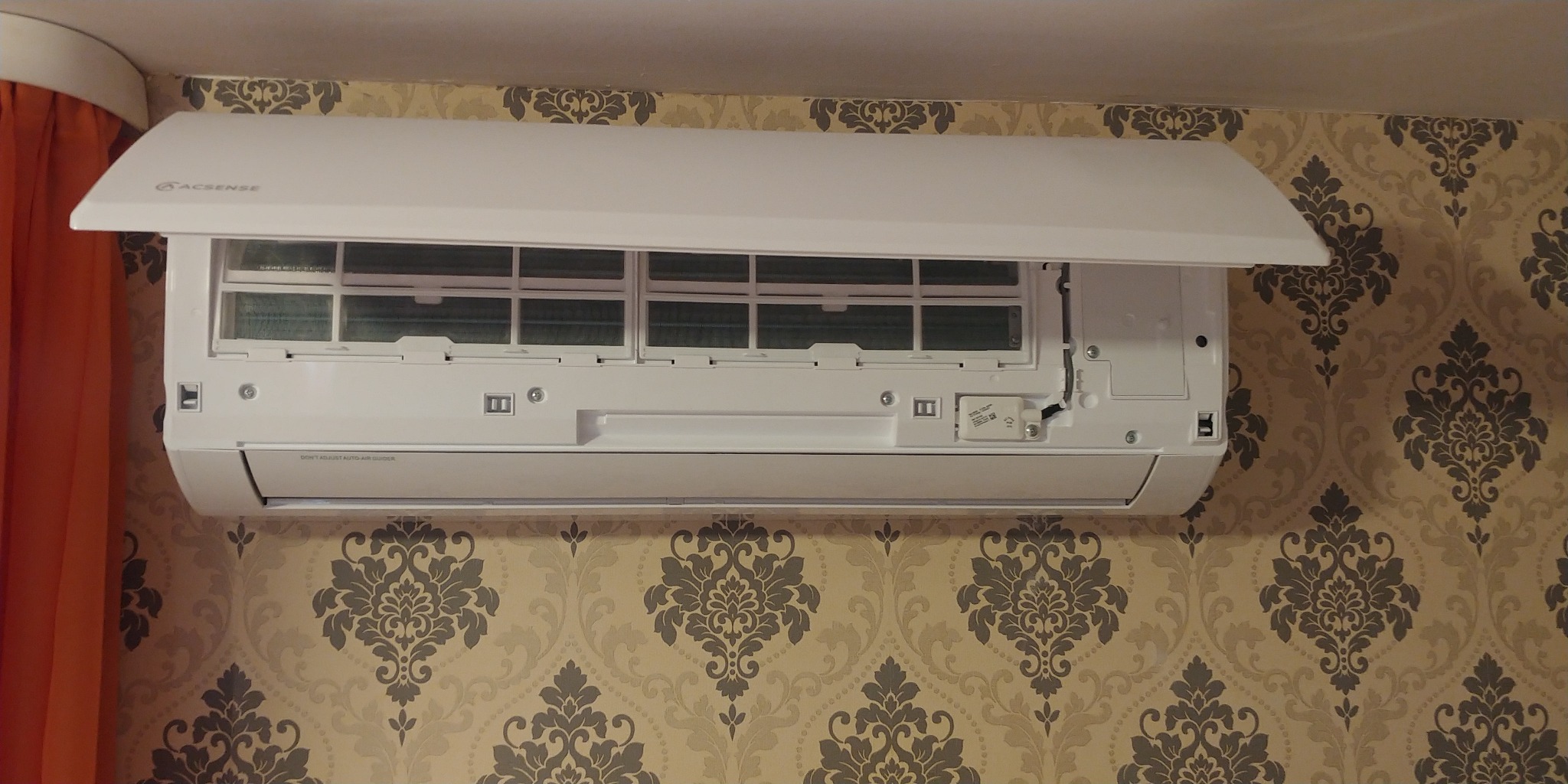 Biała jednostka wewnętrzna klimatyzatora marki Acsense z otwartą klapą, zamontowana na ścianie z tapetą w ornamenty.
