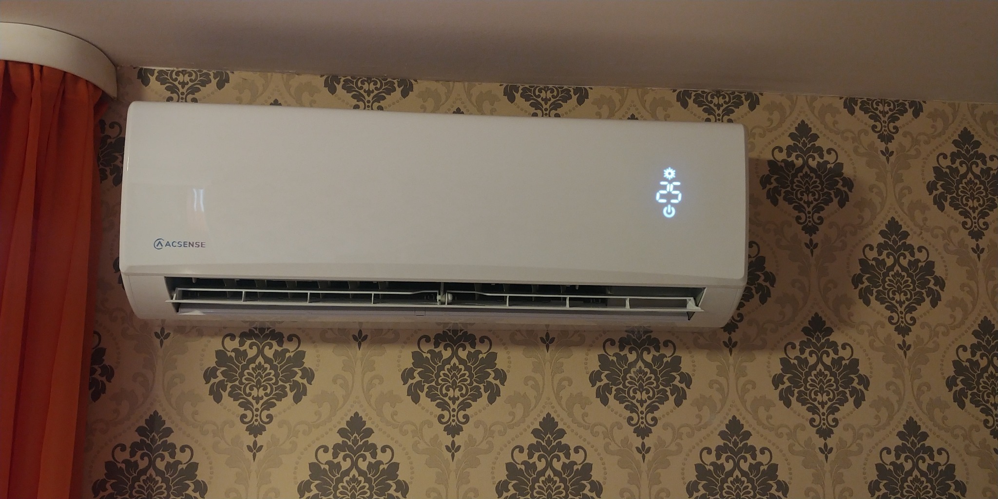 Biała klimatyzacja ścienna ACSENSE z wyświetlaczem temperatury 25 stopni Celsjusza, zamontowana na ścianie z wzorzystą tapetą w odcieniach beżu i brązu, obok pomarańczowej zasłony.