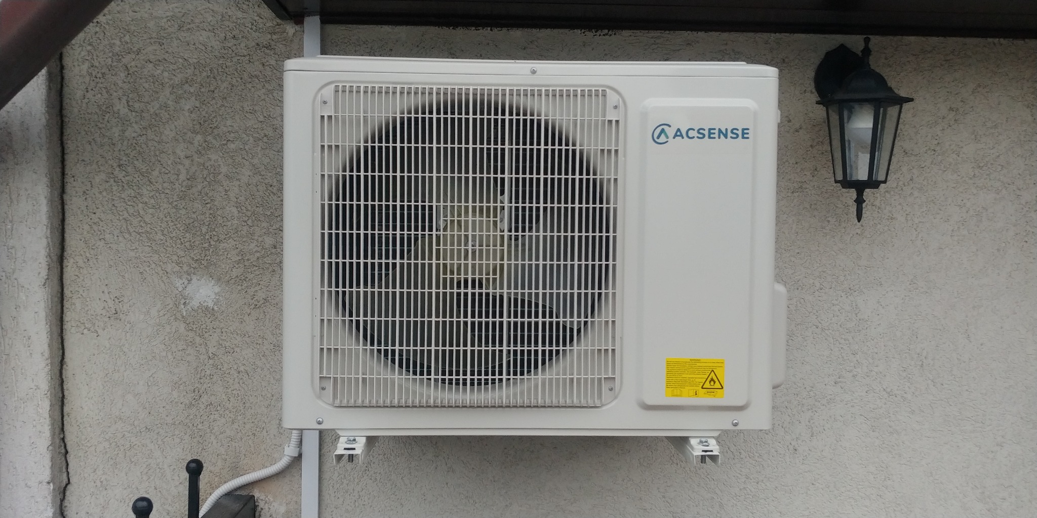 Zewnętrzna jednostka klimatyzacji marki ACSENSE zamontowana na szarej, teksturowanej ścianie budynku, obok widoczna lampa zewnętrzna.