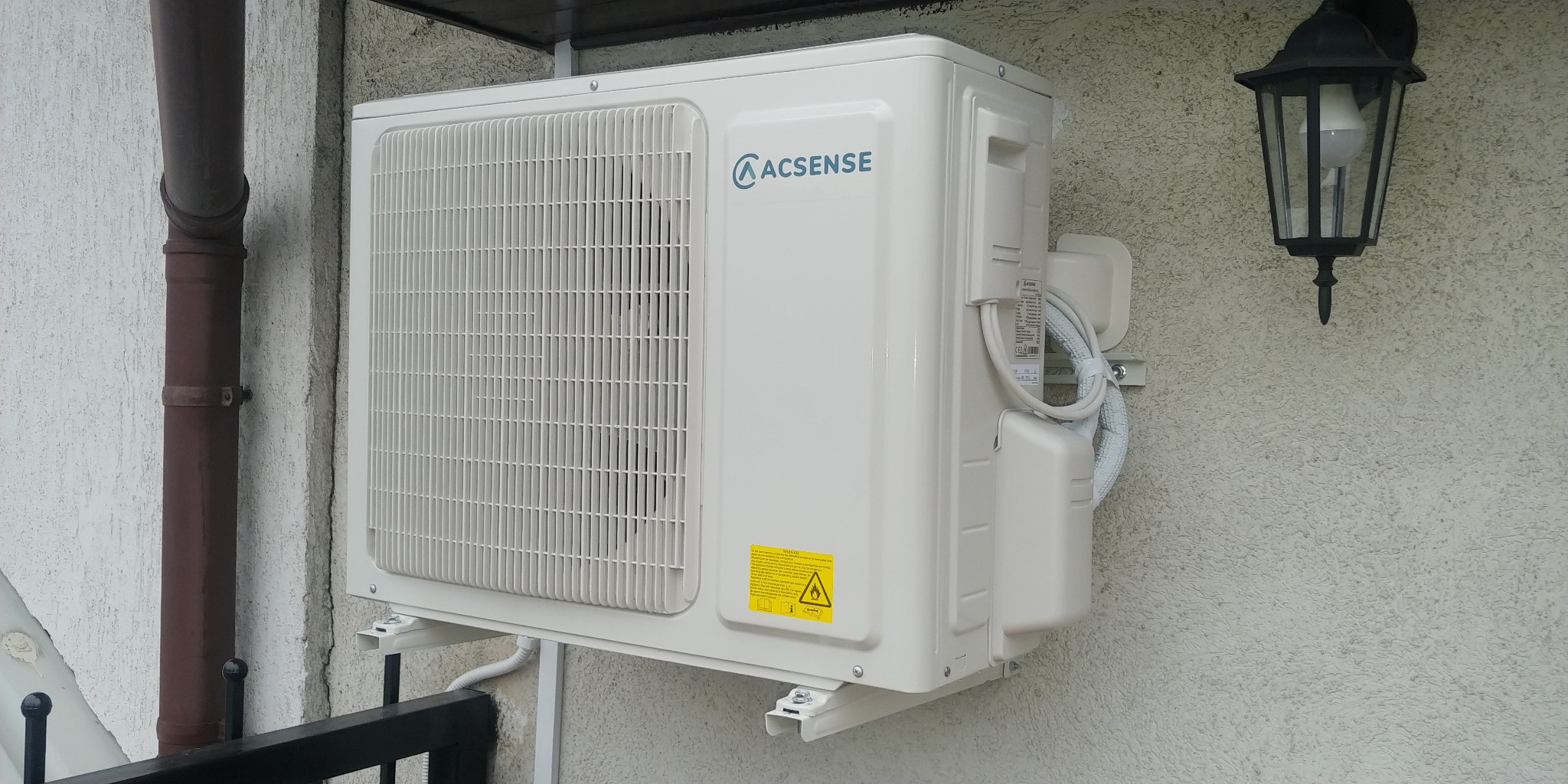 Zewnętrzna jednostka klimatyzacji marki Acsense zamontowana na elewacji budynku, widoczny brązowy rynna i czarna lampa zewnętrzna.