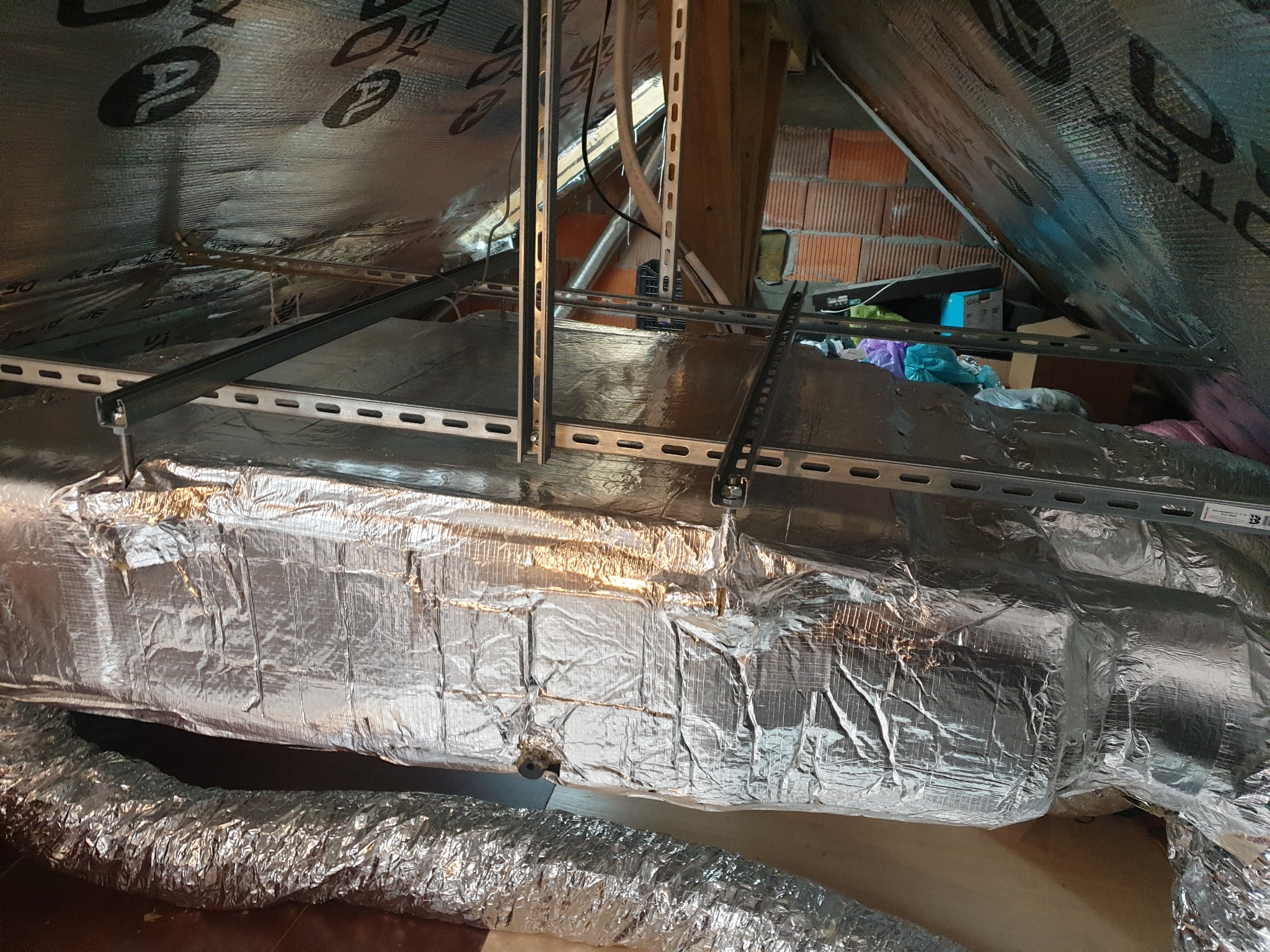Instalacja kanałów wentylacyjnych w izolowanej przestrzeni poddasza, widoczne metalowe profile montażowe i fragment ceglanej ściany.