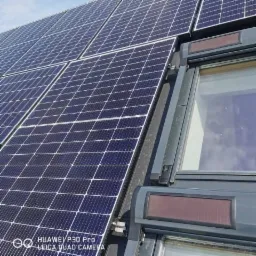 Panele słoneczne zamontowane na dachu obok okna dachowego, widoczne mocowania i konstrukcja nośna.