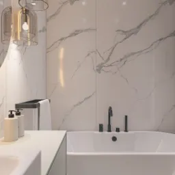 Nowoczesna łazienka z białymi marmurowymi płytkami, owalną wanną i czarną armaturą, oświetlona lampami wiszącymi z elementami złota oraz podświetlanym lustrem.