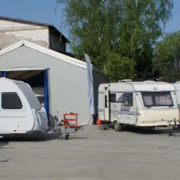 Rząd zaparkowanych przyczep kempingowych różnych marek i modeli przed szarym magazynem z otwartymi wrotami; widoczna przyczepa z czerwonym wózkiem narzędziowym.