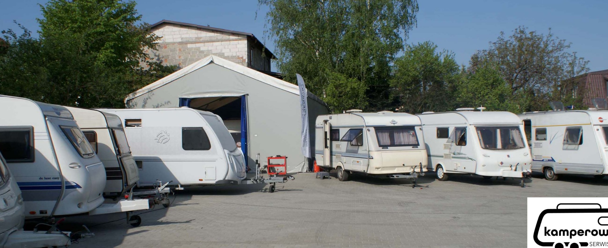 Rząd zaparkowanych przyczep kempingowych różnych marek i modeli przed szarym magazynem z otwartymi wrotami; widoczna przyczepa z czerwonym wózkiem narzędziowym.