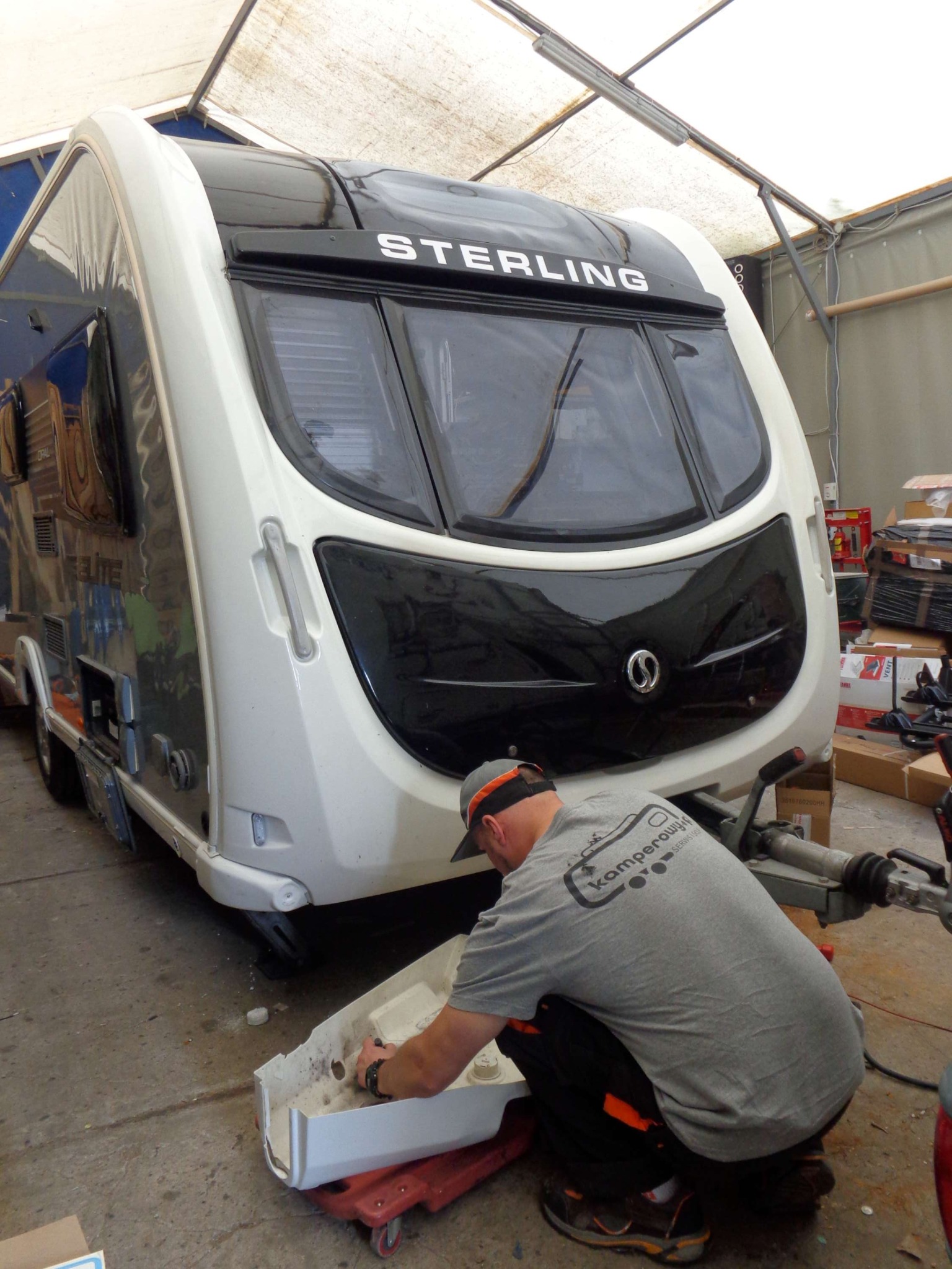 Pracownik w szarej koszulce naprawia element przyczepy kempingowej marki Sterling w warsztacie, widoczny zaczep przyczepy i kartony w tle.