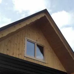 Fragment drewnianego domu w trakcie budowy z zamontowanym oknem, widoczne deski elewacyjne, czarny dach i logo firmy WINROOF na górze.