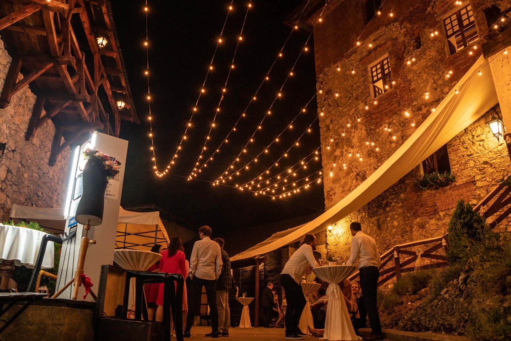 Uroczystość plenerowa nocą, oświetlona girlandami żarówek, z widoczną ścianą starego budynku z cegły i kamienia, oraz grupą osób przy stolikach koktajlowych. W lewym rogu manekin z kwiatami...