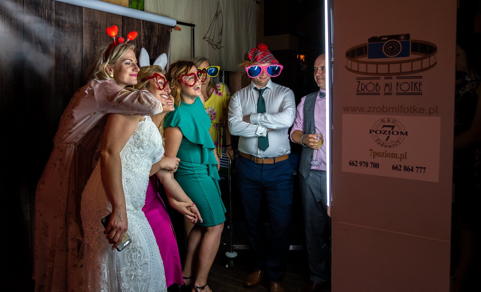 Grupa osób w przebraniach pozuje w fotobudce podczas wesela, widoczny mężczyzna z drinkiem i logo fotobudki.