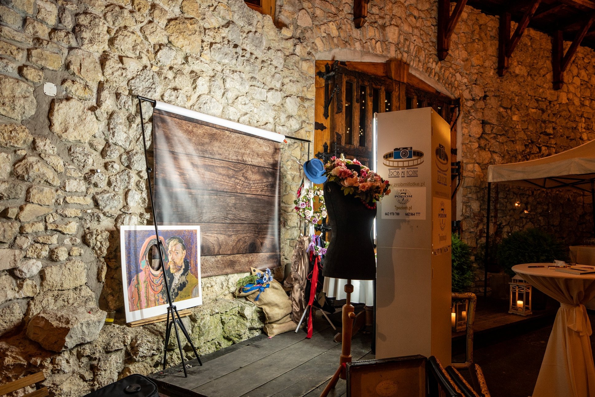 Dekoracje przy stanowisku do zdjęć z tłem imitującym drewno, manekin z kwiatami na głowie, obraz na trójnogu i kamienna ściana w tle.