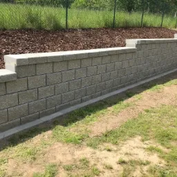 Nowo wybudowany murek oporowy z szarej kostki betonowej, oddzielający trawnik od kory drzewnej, z widoczną podmurówką i wykończeniem górnym.
