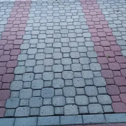 Ułożona kostka brukowa w odcieniach szarości i czerwieni, widoczne regularne wzory i fuga, z pojedynczym źdźbłem trawy wyrastającym spomiędzy czerwonych kostek.