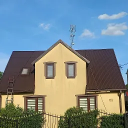 Dwupiętrowy dom z brązowym dachem z blachy falistej, drabina oparta o dach, żółta elewacja z numerem 53, antena na dachu, ogrodzenie z ozdobnymi grotami i krzewami, błękitne niebo z chmurami.