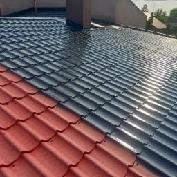 Częściowo pomalowany dach z blachodachówki w kolorze czerwonym i grafitowym, widoczny komin i sąsiednie budynki.