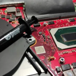 Otwarty laptop z widoczną czerwoną płytą główną, procesorem, wentylatorem i tubką pasty termoprzewodzącej Thermal Grizzly, gotową do nałożenia.