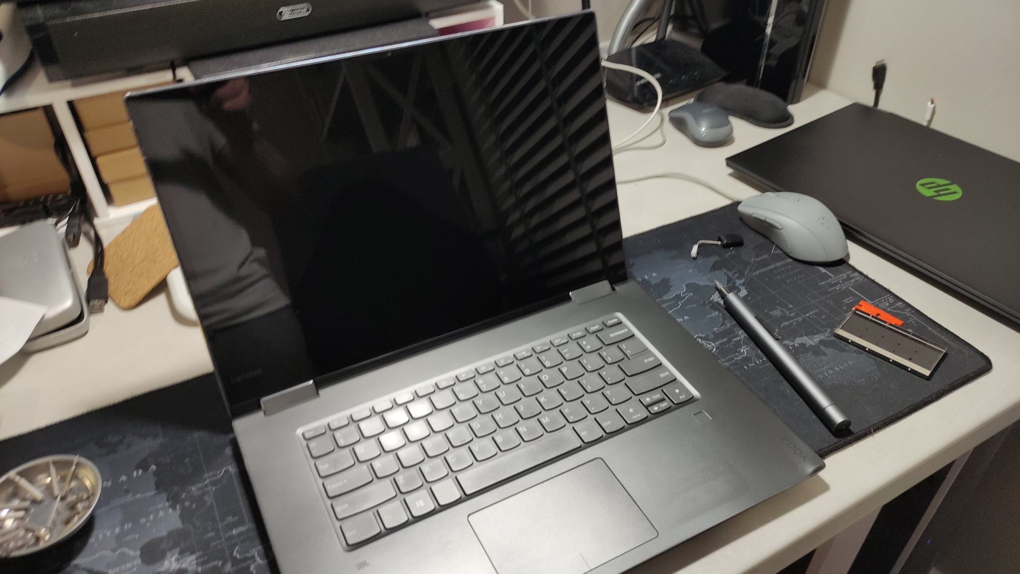 Laptop Lenovo Yoga z czarnym ekranem, obok narzędzia do naprawy, pad z mapą świata i mysz komputerowa, drugi laptop HP w tle.
