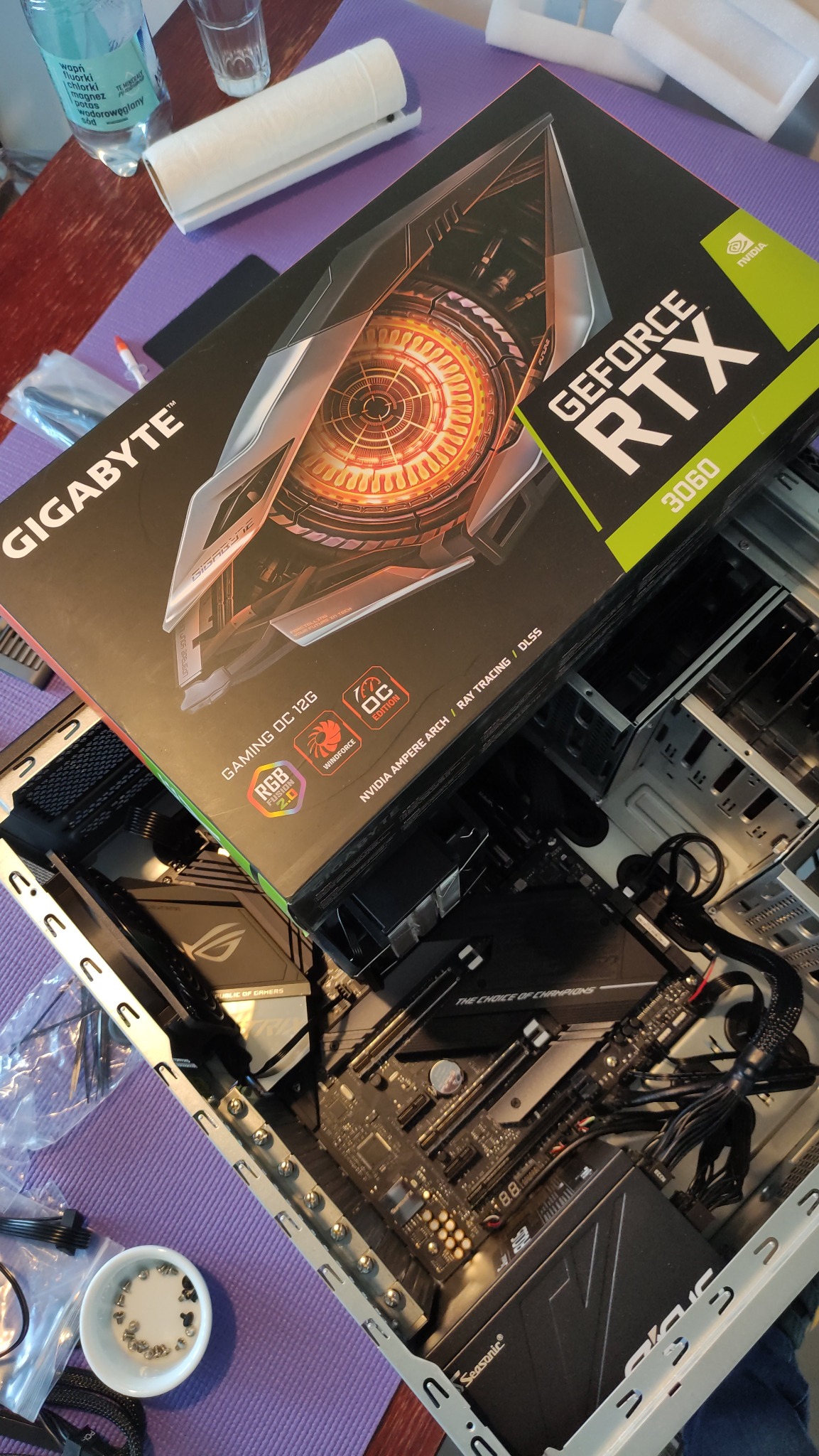 Pudełko karty graficznej Gigabyte GeForce RTX 3060 leżące na otwartej obudowie komputera stacjonarnego z widoczną płytą główną, zasilaczem Seasonic i drobnymi elementami montażowymi, sugerujące...