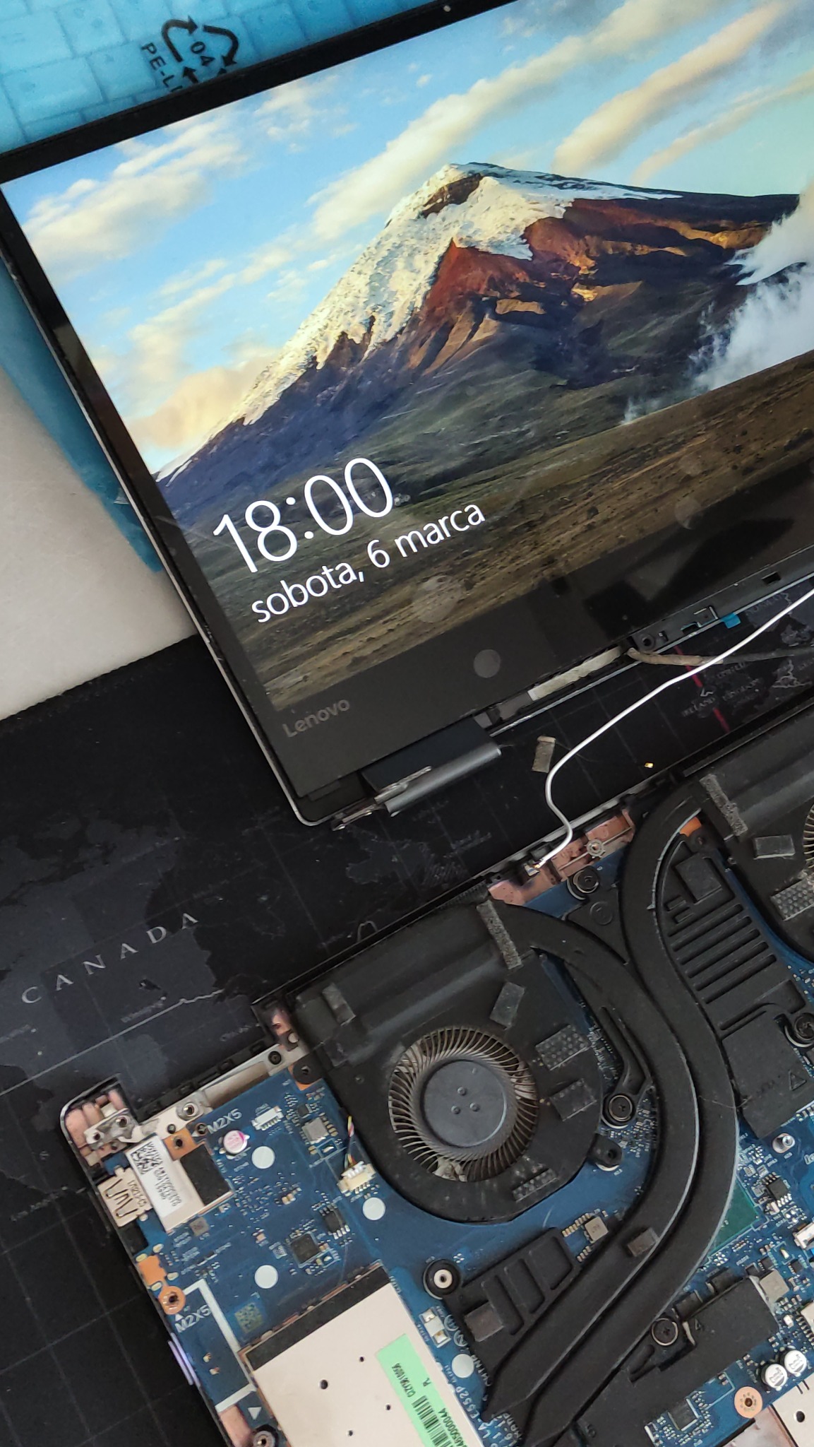 Otwarty laptop Lenovo podczas naprawy, widoczna płyta główna, wentylator chłodzenia i ekran z wyświetloną godziną 18:00 oraz datą sobota, 6 marca, na tle górskiego krajobrazu.