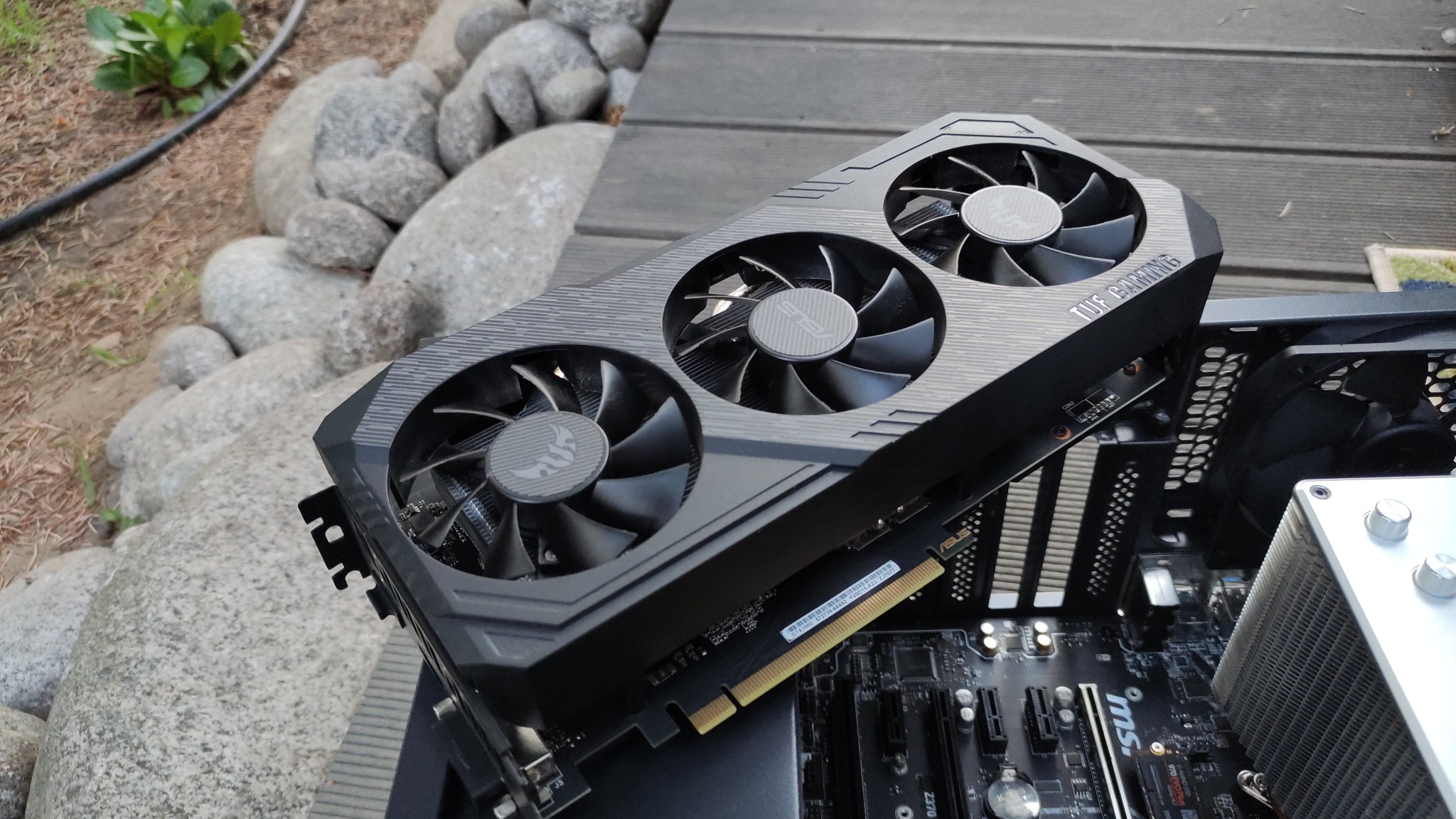 Karta graficzna ASUS TUF Gaming z trzema wentylatorami umieszczona na płycie głównej wewnątrz obudowy komputera, na tle drewnianego podestu i kamieni.