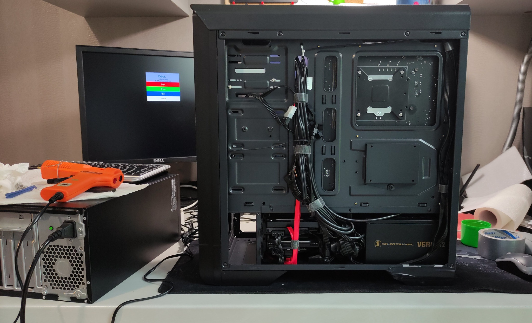 Wnętrze otwartej obudowy komputera stacjonarnego z widocznymi kablami, zasilaczem SilentiumPC Vero L2 oraz drugim komputerem z podłączonym monitorem Dell w tle, na którym wyświetlany jest test...