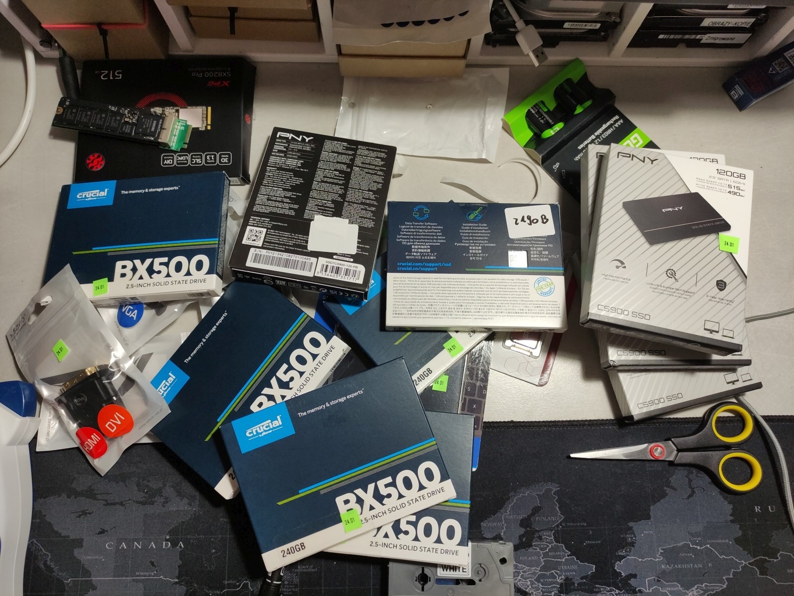 Stół z rozłożonymi pudełkami dysków SSD Crucial BX500 i PNY CS900 o różnych pojemnościach, adapterami HDMI/DVI, nożyczkami i pudełkiem dysku XPG SX8200 Pro 512GB na podkładce z mapą świata.