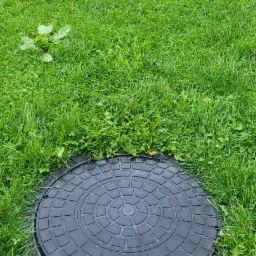 Czarna pokrywa studni z wzorem kołowym, umieszczona w trawie, z widocznym niebieskim wężem na skraju.