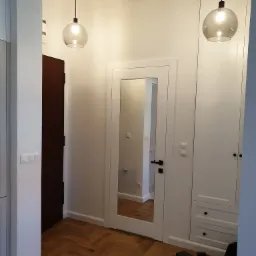 Nowoczesny przedpokój z parkietem w jodełkę, białymi drzwiami z szybą i zabudowaną szafą, oświetlony dwoma szklanymi lampami wiszącymi; na podłodze widoczna wkrętarka i pudełko narzędzi.