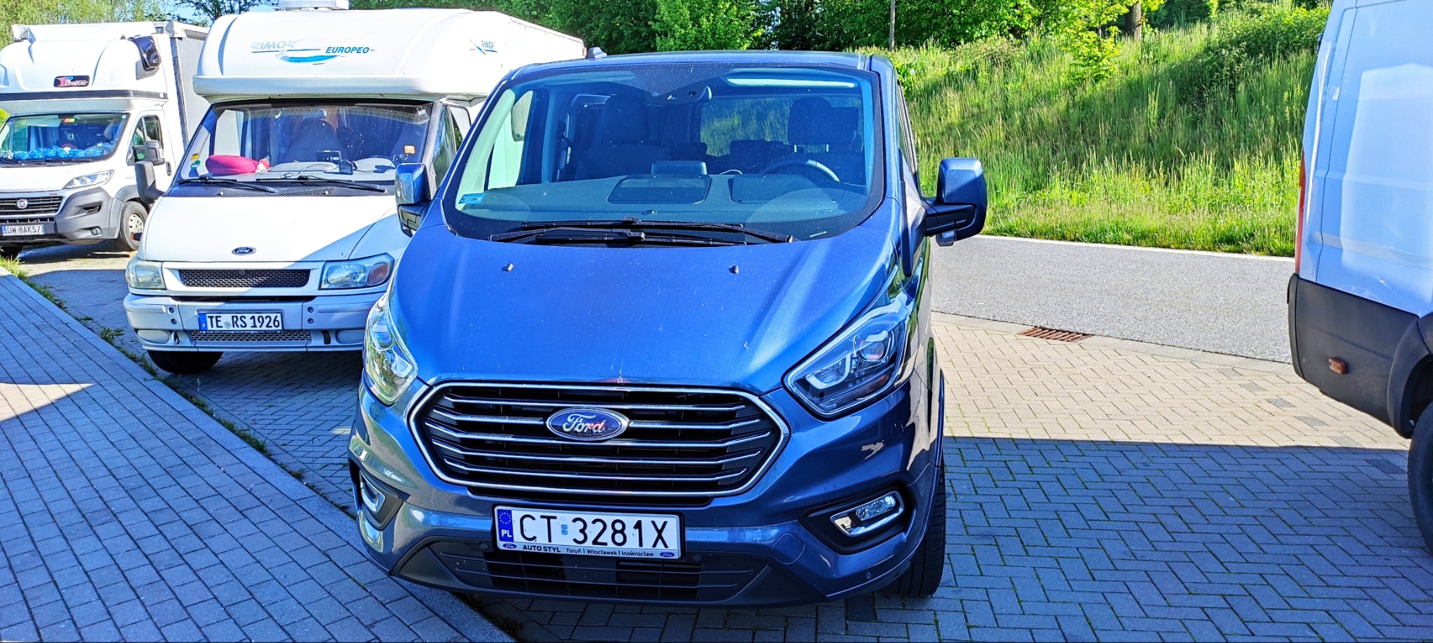 Niebieski Ford Transit z polską tablicą rejestracyjną CT 3281X zaparkowany na brukowanym parkingu, z widocznymi kamperami w tle.