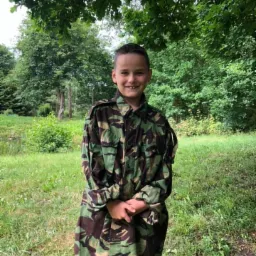 Uśmiechnięty chłopiec w pełnym stroju moro na tle leśnej zieleni, prawdopodobnie na obozie.